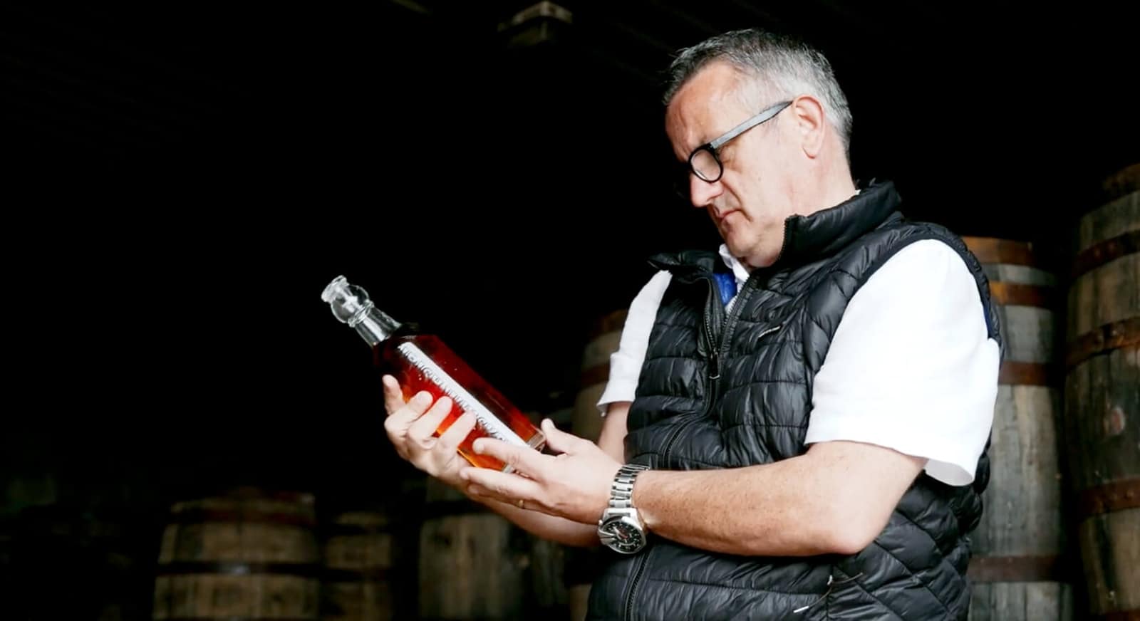 Man examining a bottle in a wine cellar with barrels in the background