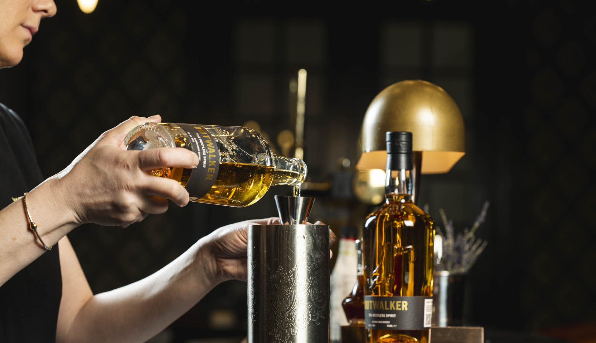 Person pouring whiskey into a glass with a bottle of Outwalker whiskey in the background.